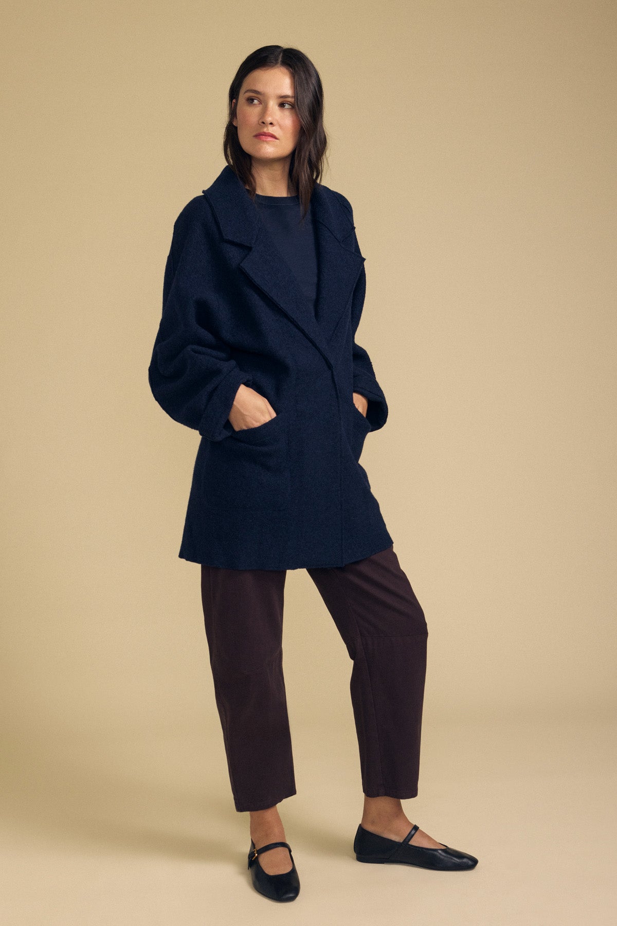 A woman stands against a beige background, wearing Velvet by Graham & Spencer's ABBY BLAZER styled loosely over a dark shirt, paired with brown pants and black flat shoes, her hands tucked in the blazer pockets as she looks to the side—a perfect example of everyday layering.-41834056908993
