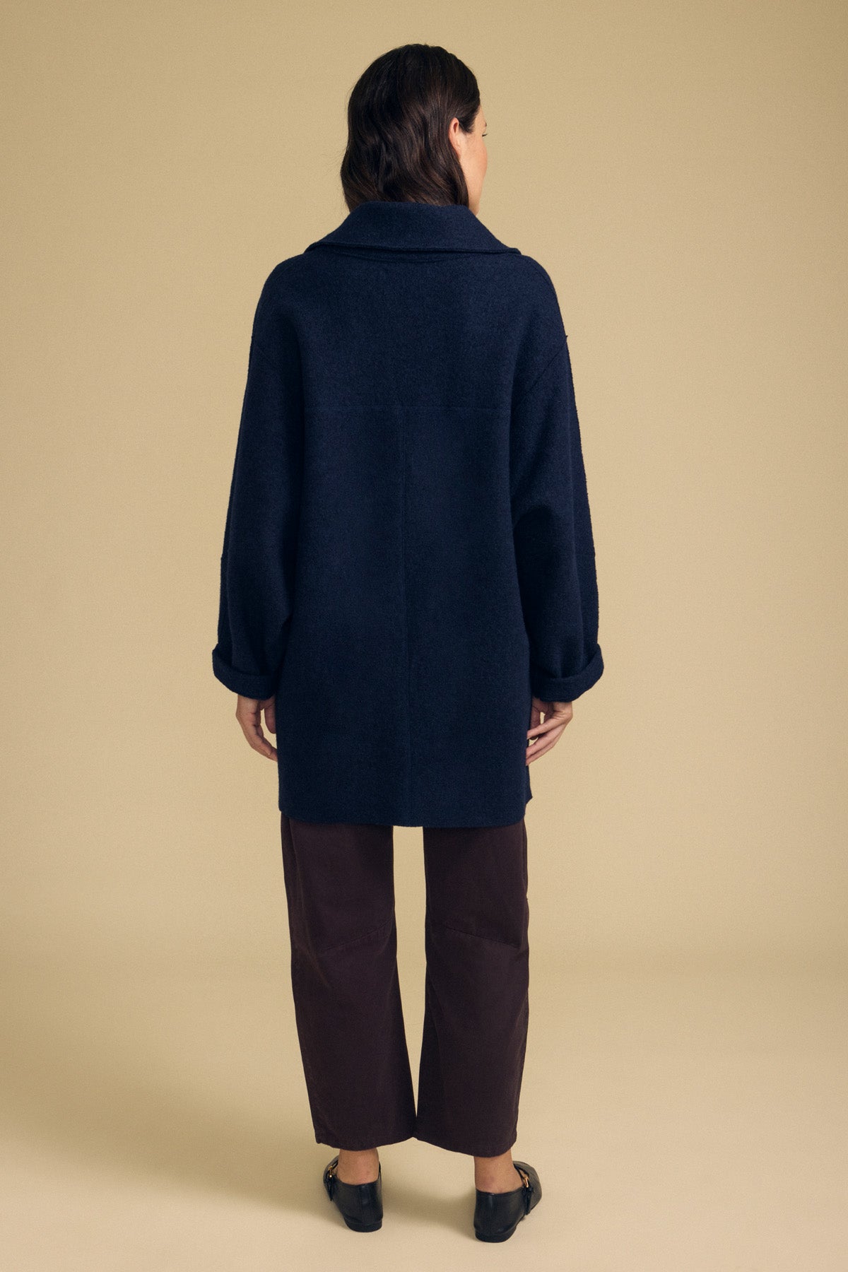 A person stands facing away, wearing the ABBY BLAZER by Velvet by Graham & Spencer in dark navy wool, paired with brown pants and black shoes against a beige background—perfect for everyday layering.