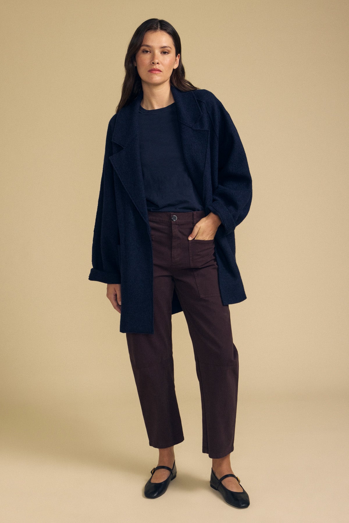 A person stands against a beige background wearing the ABBY BLAZER by Velvet by Graham & Spencer layered over a black shirt, paired with brown pants and black shoes, showcasing effortless everyday layering.