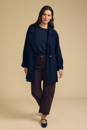 A person stands against a beige background wearing the ABBY BLAZER by Velvet by Graham & Spencer for everyday layering, paired with a navy top, brown wide-leg pants, and black Mary Jane shoes.