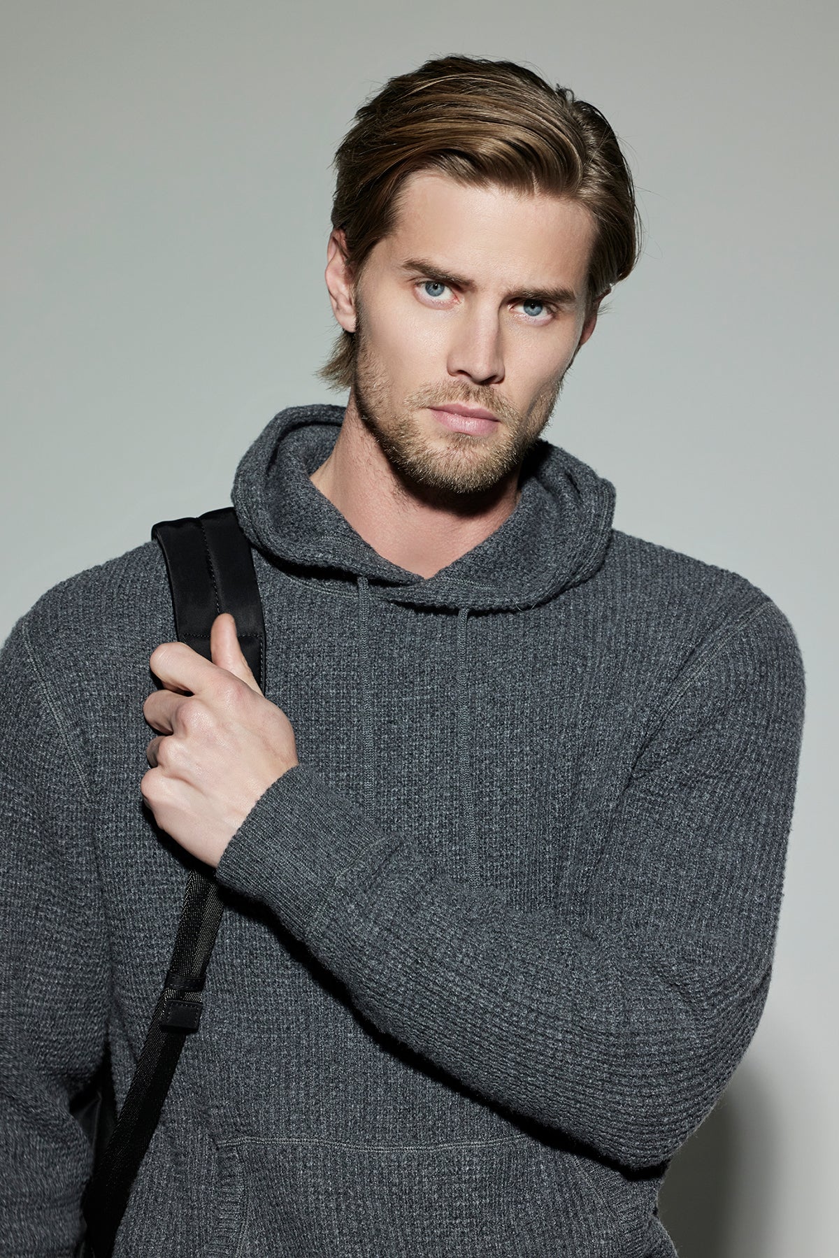   A man with light brown hair and a beard wears the SHANE SWEATER HOODIE in gray, from Velvet by Graham & Spencer, holding a black strap over his shoulder, and stands against a plain background. 