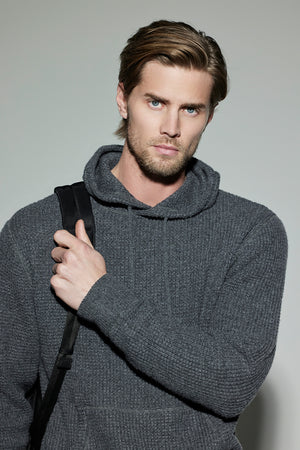 A man with light brown hair and a beard wears the SHANE SWEATER HOODIE in gray, from Velvet by Graham & Spencer, holding a black strap over his shoulder, and stands against a plain background.