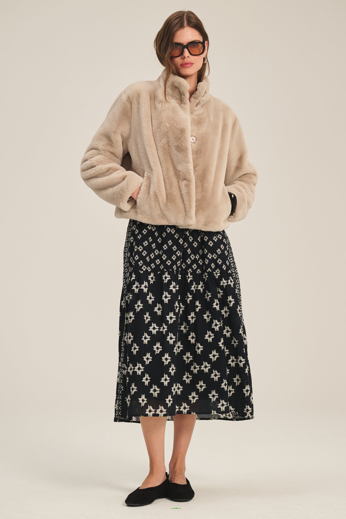   A woman wearing the ELEN DRESS by Velvet by Graham & Spencer, paired with a tan faux fur jacket, black shoes, and large sunglasses, stands against a plain background. 