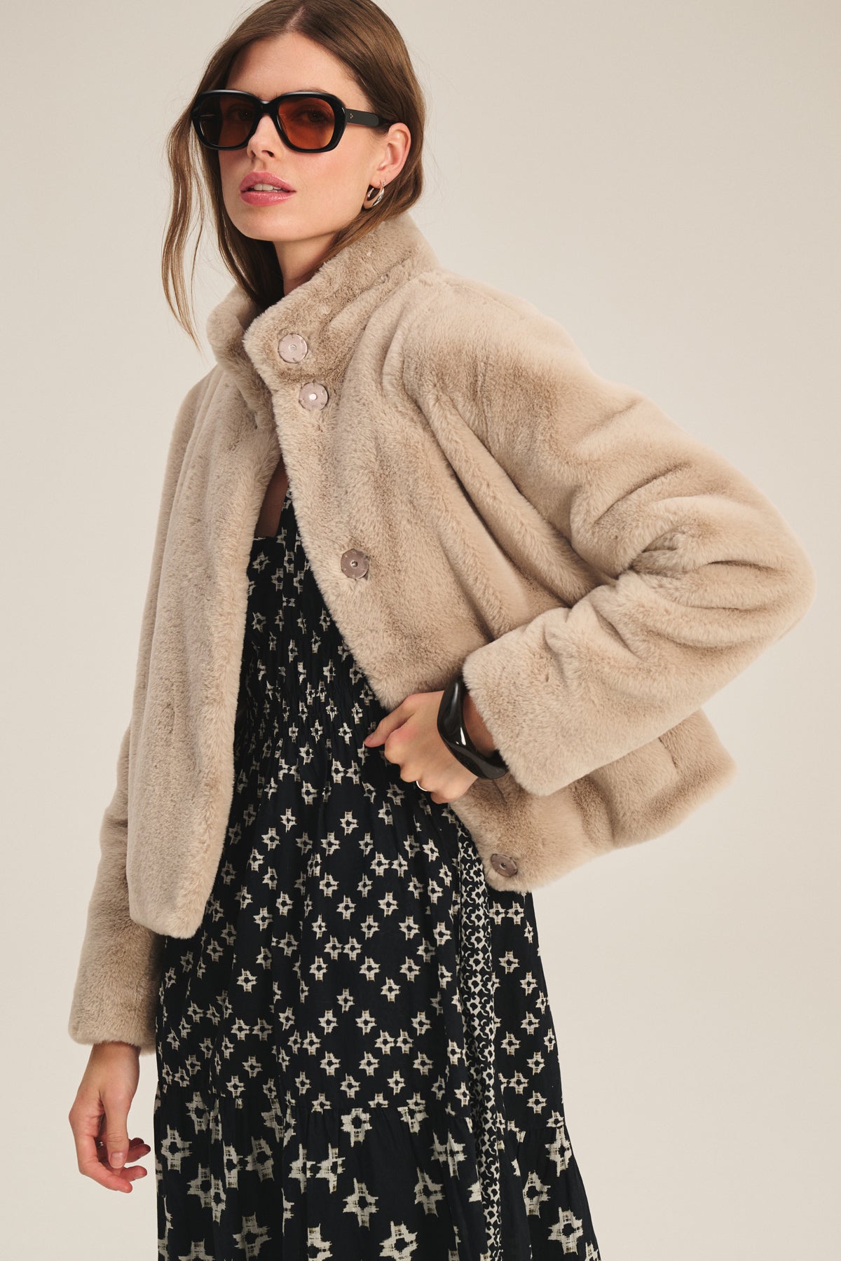 A woman models the ELEN DRESS by Velvet by Graham & Spencer—a black patterned puff sleeve dress—styled with sunglasses and a beige faux fur jacket against a plain background.-39815218168001