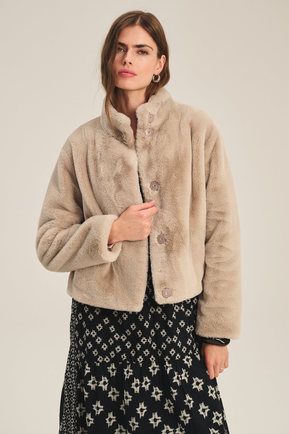 A woman models the SHEENA LUXE FAUX FUR JACKET by Velvet by Graham & Spencer in light beige with a mock neck, layered over a black and white patterned dress, standing against a plain background.-39815218135233