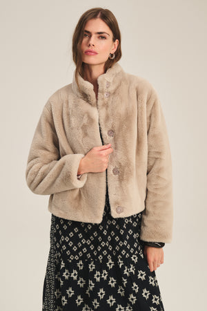 A woman models the SHEENA LUXE FAUX FUR JACKET by Velvet by Graham & Spencer in light beige with a mock neck, layered over a black and white patterned dress, standing against a plain background.