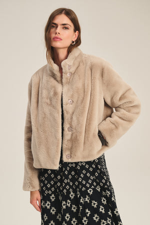 A woman wears the SHEENA LUXE FAUX FUR JACKET by Velvet by Graham & Spencer—a beige, mock-neck piece—over a black dress with a white star pattern, standing against a plain background.