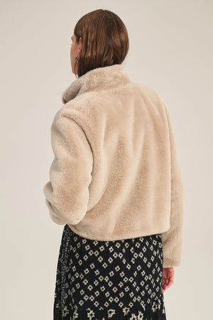 A person stands facing away in the SHEENA LUXE FAUX FUR JACKET by Velvet by Graham & Spencer, featuring a beige faux fur design with a mock neck collar, paired with a black skirt with a white geometric pattern against a plain background.