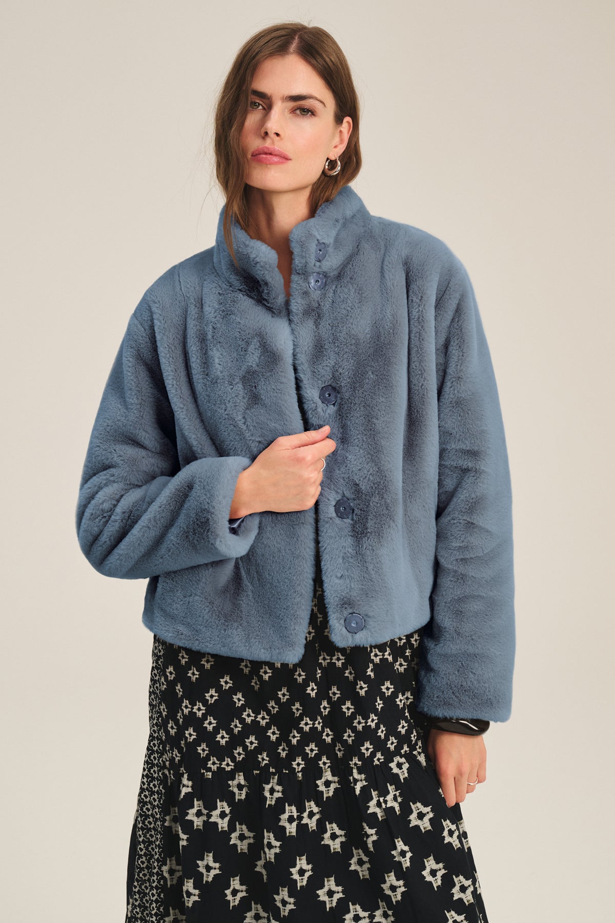 A woman models the SHEENA LUXE FAUX FUR JACKET by Velvet by Graham & Spencer in blue, styled over a black and white patterned dress against a simple light background.-39839681085633