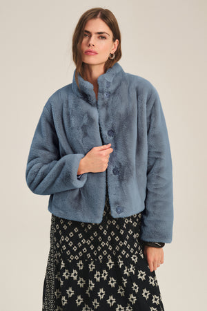 A woman models the SHEENA LUXE FAUX FUR JACKET by Velvet by Graham & Spencer in blue, styled over a black and white patterned dress against a simple light background.