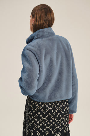 Model faces away from the camera, wearing the SHEENA LUXE FAUX FUR JACKET by Velvet by Graham & Spencer in light blue and a black skirt with white geometric print—a cozy, stylish look for chilly days.