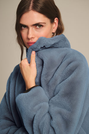 Model in the SHEENA LUXE FAUX FUR JACKET by Velvet by Graham & Spencer, light blue, cozy fit, holding the collar up near her face against a neutral background.