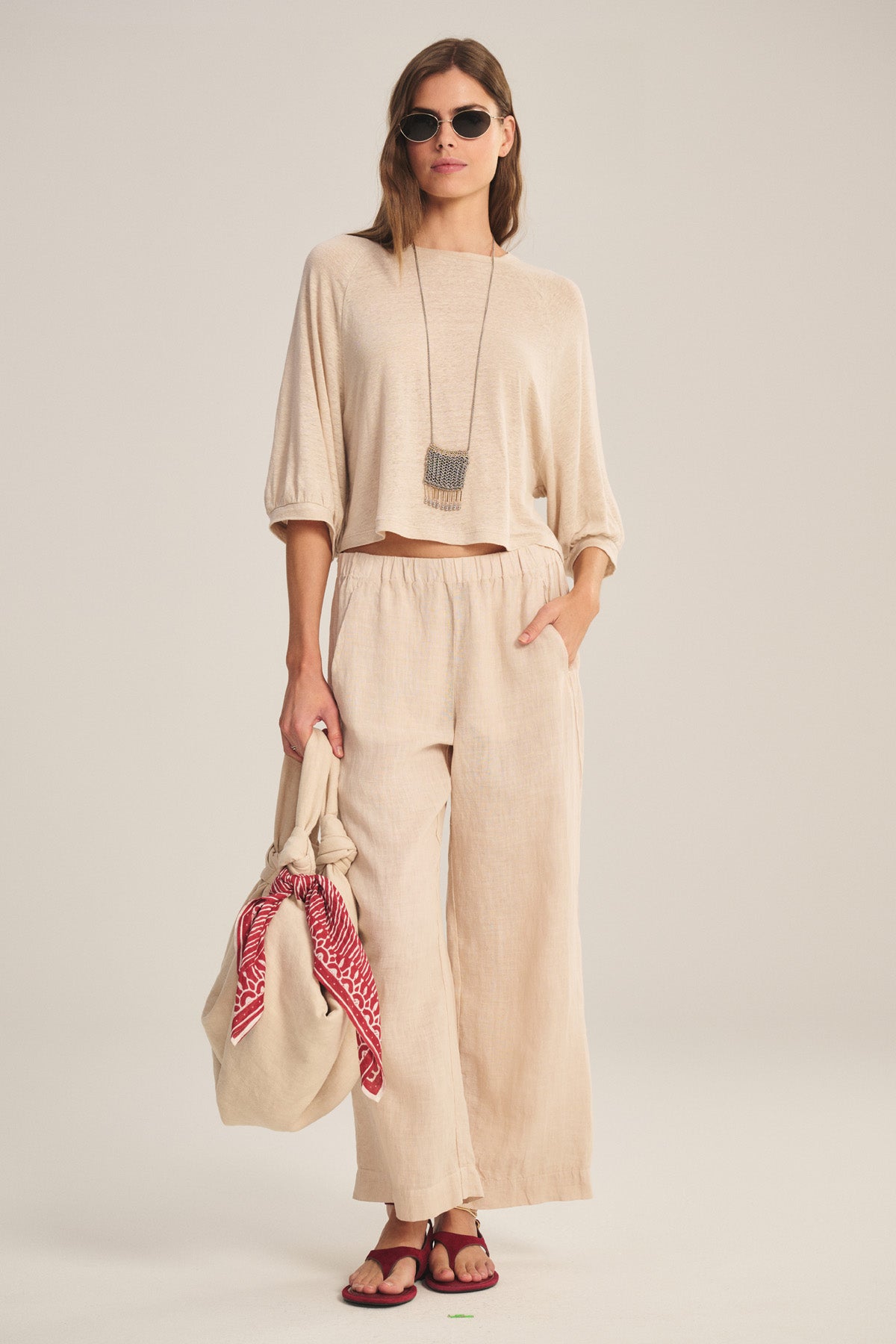 A woman styles the Velvet by Graham & Spencer LOLA LINEN PANT with a loose beige top, sunglasses, sandals, a long necklace, and carries a beige bag accented by a red patterned scarf.