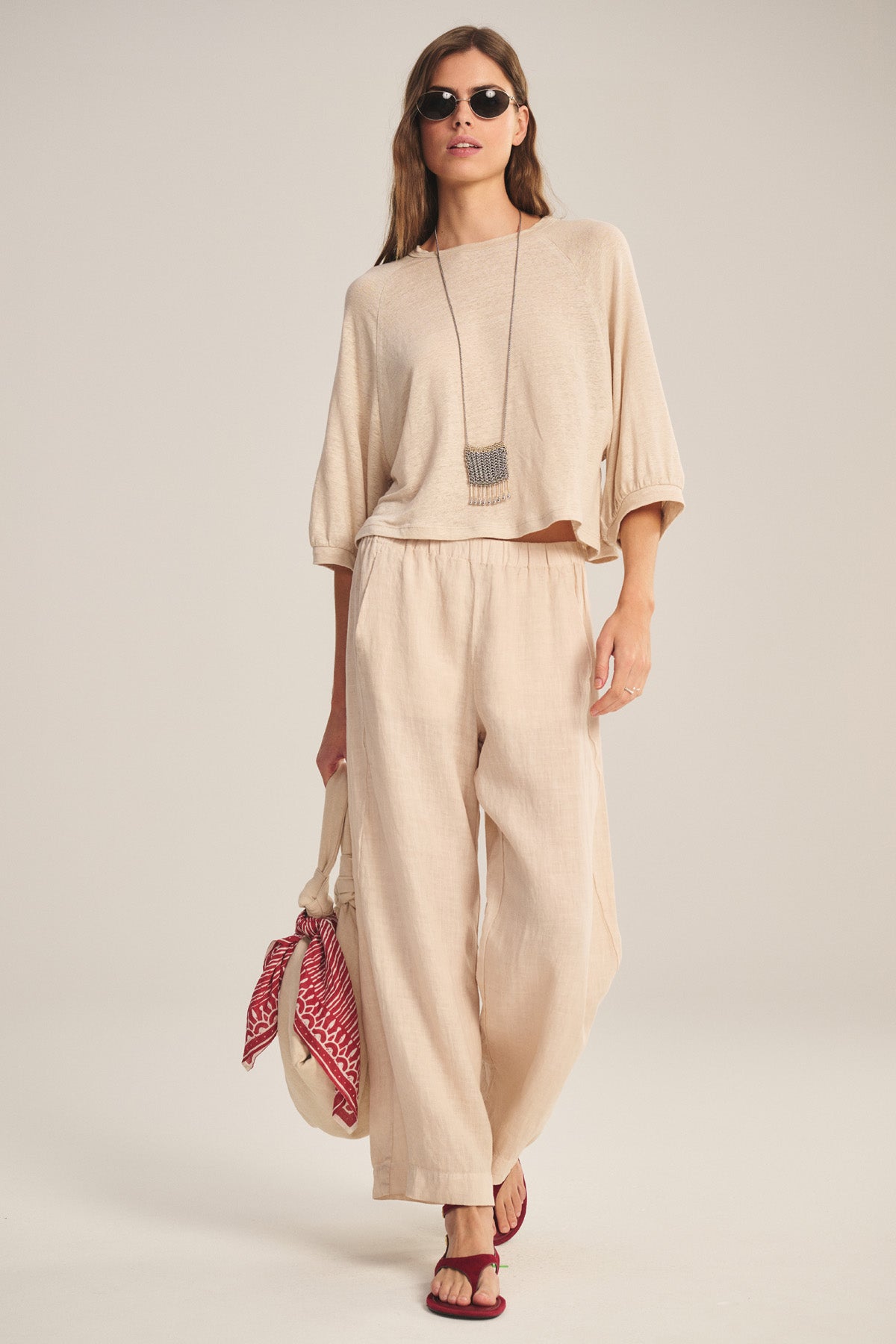 Wearing Velvet by Graham & Spencer’s LOLA LINEN PANT with an elastic waist and a matching top, carrying a tote bag with a red patterned scarf, sunglasses, long necklace, and red sandals.-40909840711873
