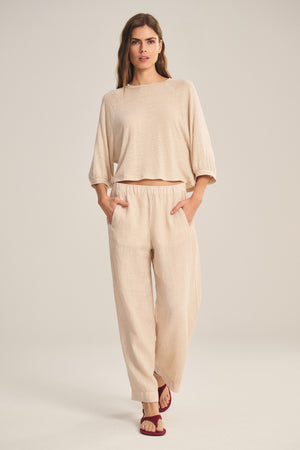 A woman stands facing forward wearing the Velvet by Graham & Spencer LOLA LINEN PANT, a lightweight linen trouser with an elastic waist, paired with a loose beige long-sleeve top and red sandals against a plain background.