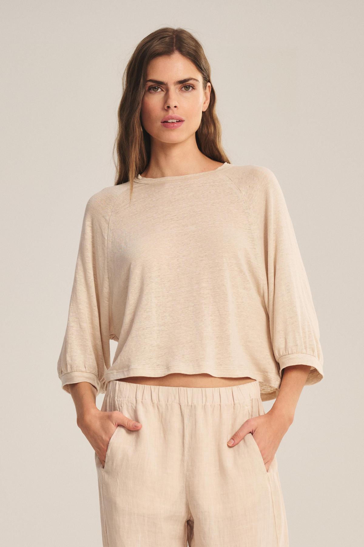 A woman faces forward against a plain background, wearing the SHELLY TEE by Velvet by Graham & Spencer—a breathable beige linen knit top with a cropped silhouette—paired with matching pants and her hands in her pockets.
