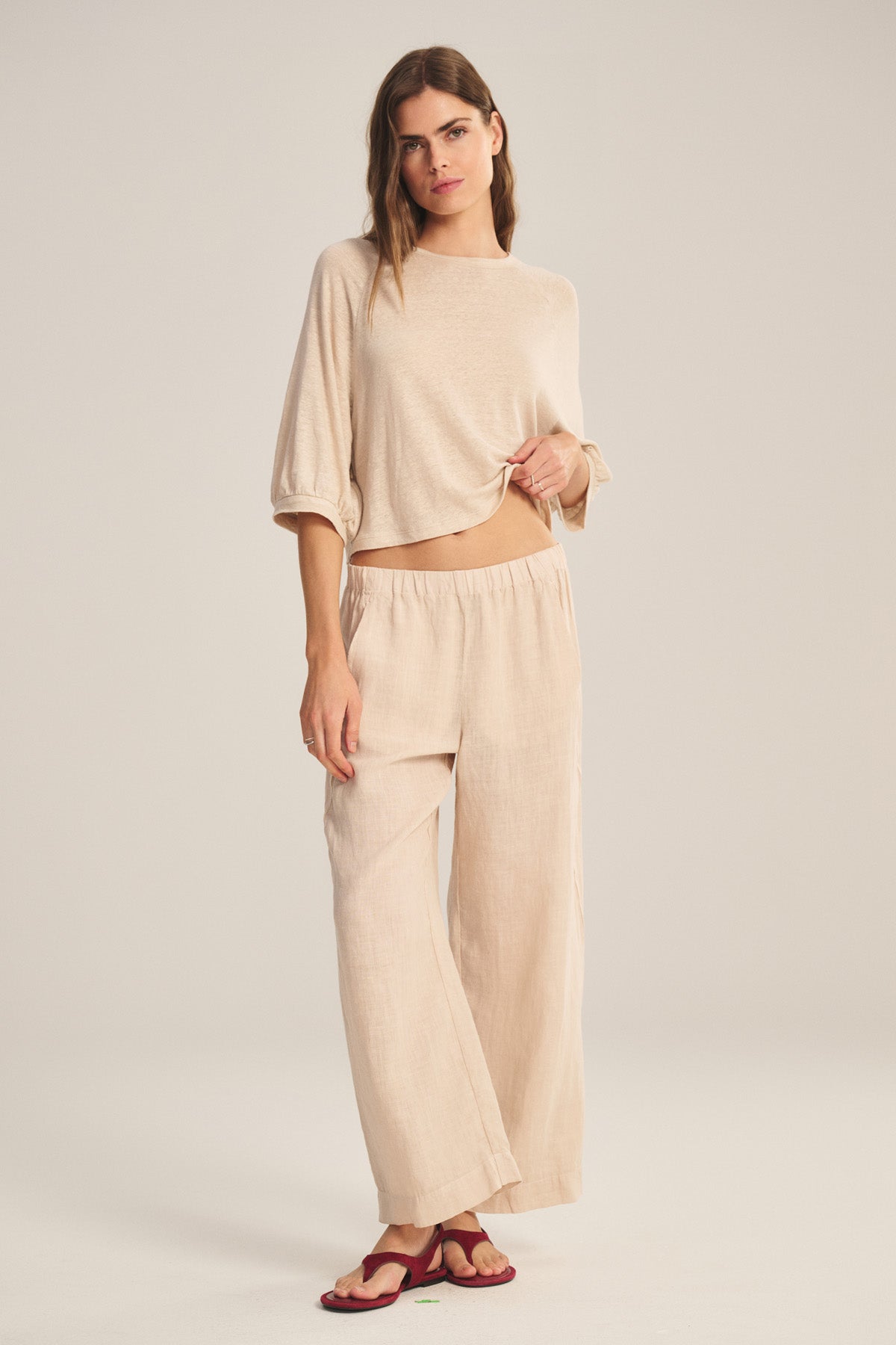 A woman stands against a plain background wearing Velvet by Graham & Spencer's LOLA LINEN PANT—a lightweight linen, elastic waist pull-on trouser—paired with a loose beige top and red sandals, lifting her top to reveal the stylish pants.