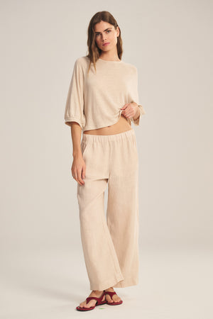 A woman stands against a plain background wearing Velvet by Graham & Spencer's LOLA LINEN PANT—a lightweight linen, elastic waist pull-on trouser—paired with a loose beige top and red sandals, lifting her top to reveal the stylish pants.