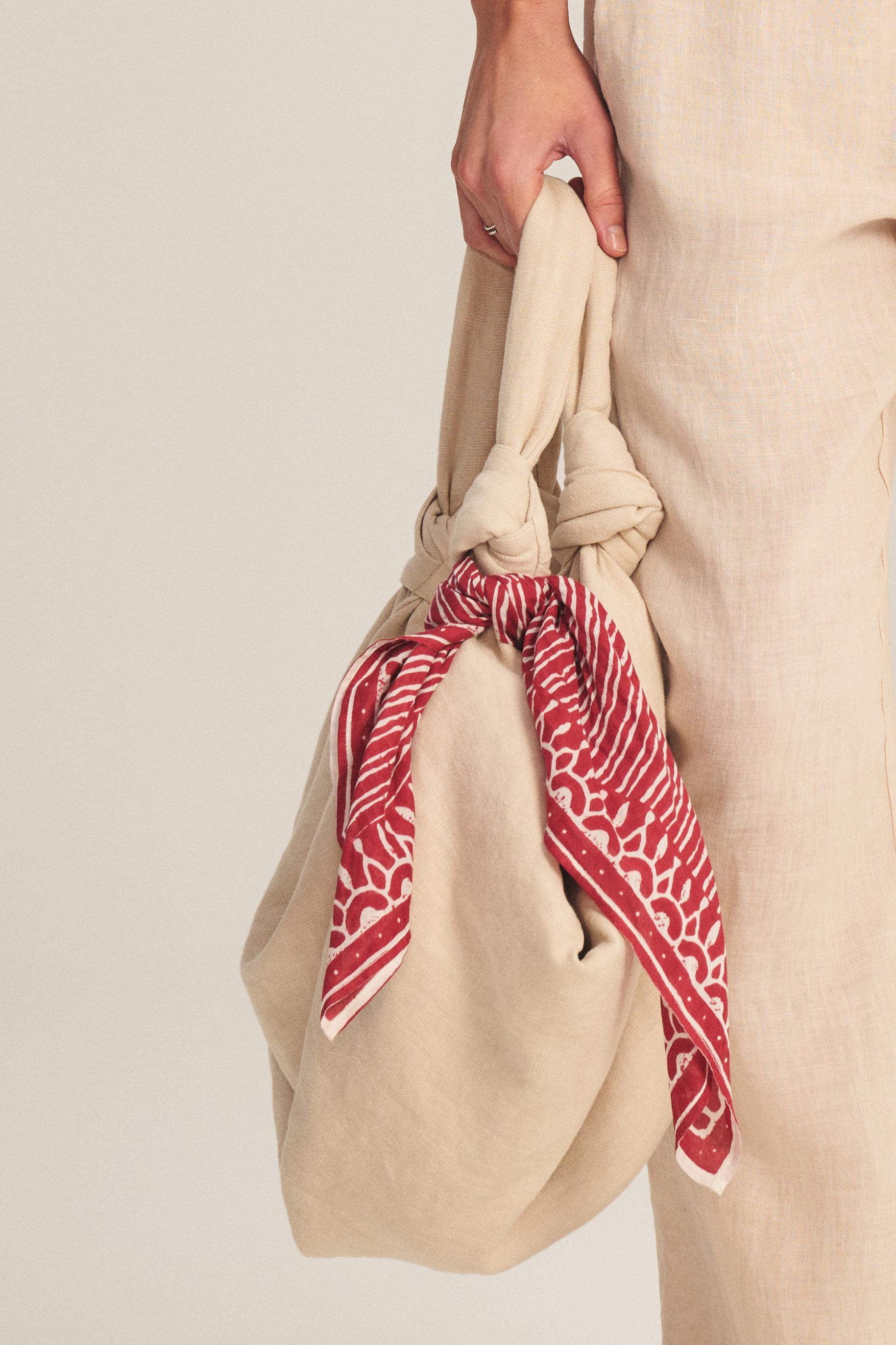 A person holds a beige fabric bag with knotted handles, accessorized with the LISETTE SCARF by Velvet by Graham & Spencer—a red patterned, lightweight cotton scarf—while dressed in beige linen clothing.