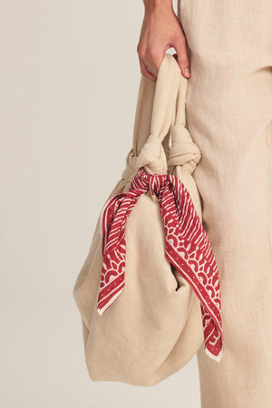 A person holds a beige fabric bag with knotted handles, accessorized with the LISETTE SCARF by Velvet by Graham & Spencer—a red patterned, lightweight cotton scarf—while dressed in beige linen clothing.