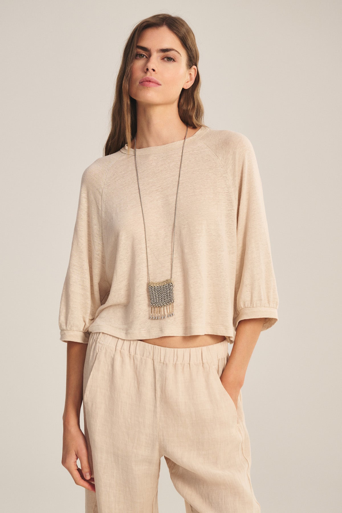 A woman wears the SHELLY TEE by Velvet by Graham & Spencer with matching pants, accessorized with a long pendant necklace, standing with one hand in her pocket against a neutral background.