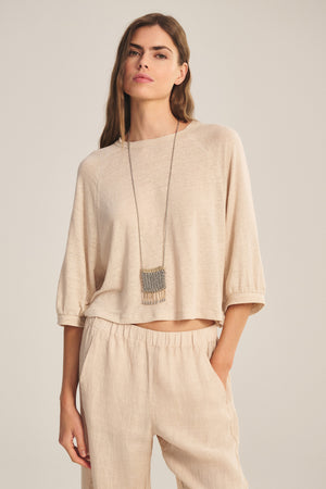 A woman wears the SHELLY TEE by Velvet by Graham & Spencer with matching pants, accessorized with a long pendant necklace, standing with one hand in her pocket against a neutral background.