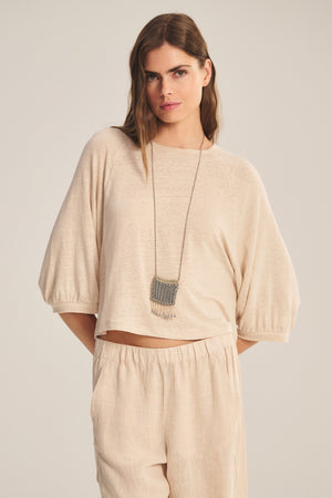 A woman wears the SHELLY TEE by Velvet by Graham & Spencer—a loose, beige linen knit top with relaxed raglan sleeves—paired with matching pants and a long necklace, standing against a neutral background.