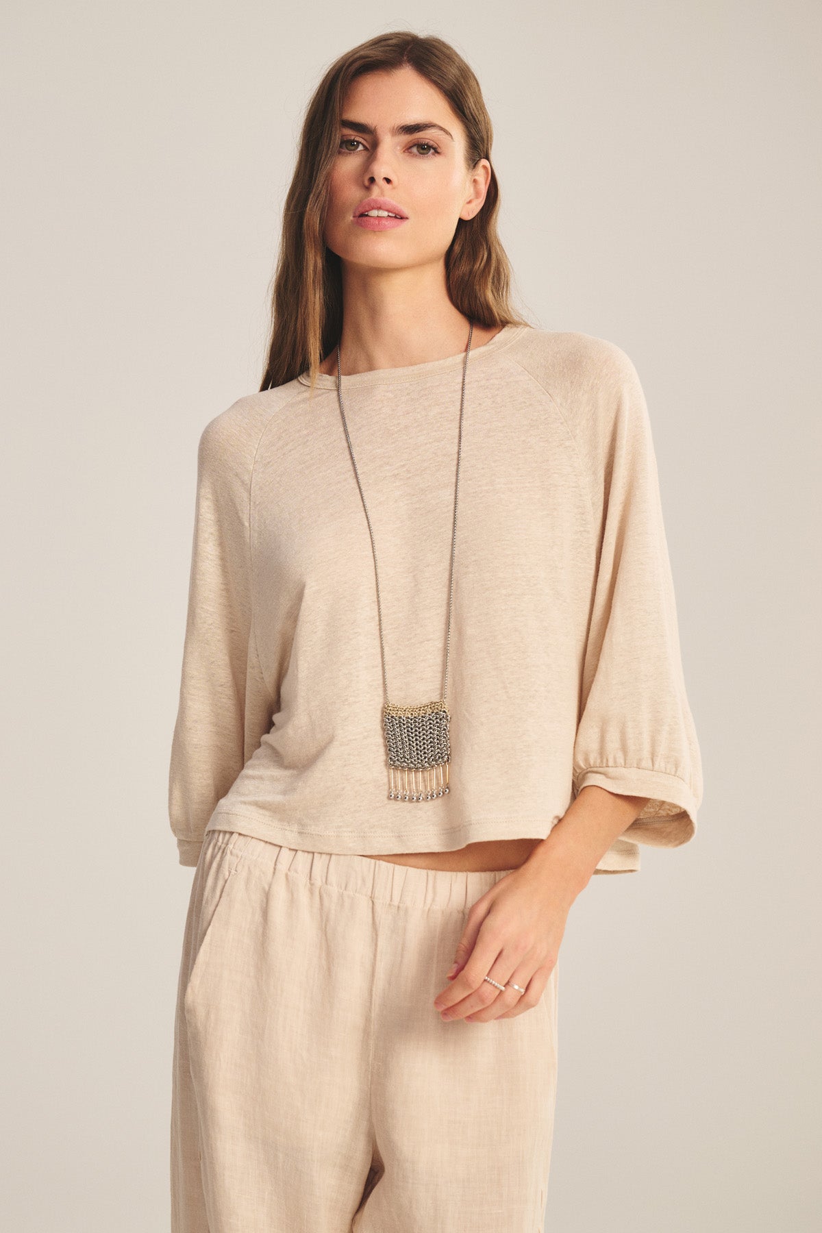 A person wearing the SHELLY TEE by Velvet by Graham & Spencer, a loose beige long-sleeve top with relaxed raglan sleeves and matching pants, accessorized with a long necklace, stands against a plain background.-40910148337857