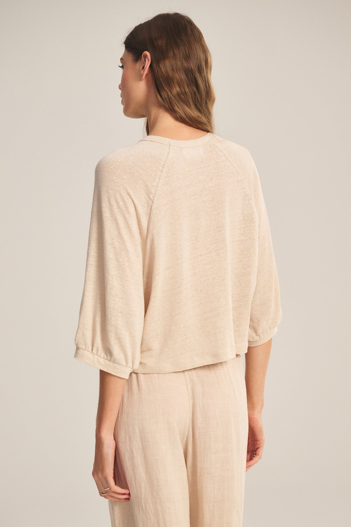A woman faces away, wearing the SHELLY TEE by Velvet by Graham & Spencer—a light beige, breathable linen knit top—paired with loose-fitting pants against a plain background.-40910148305089