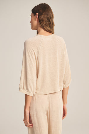 A woman faces away, wearing the SHELLY TEE by Velvet by Graham & Spencer—a light beige, breathable linen knit top—paired with loose-fitting pants against a plain background.