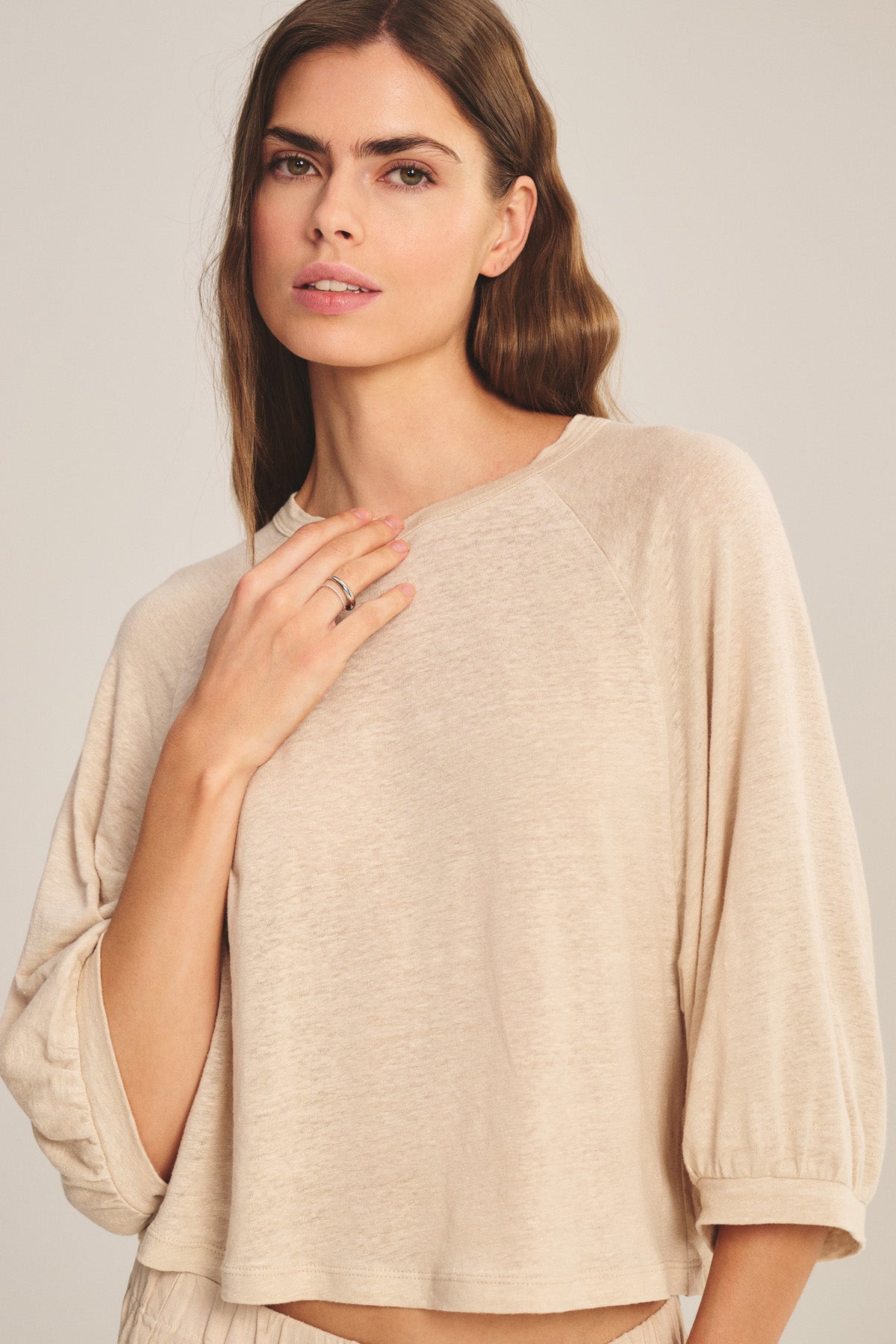 A woman with long brown hair models the SHELLY TEE by Velvet by Graham & Spencer—a cropped linen top in light beige—standing against a neutral background and looking at the camera.-40910148272321