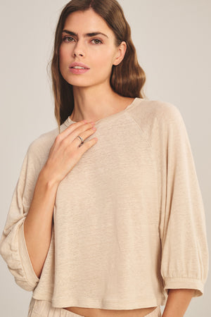 A woman with long brown hair models the SHELLY TEE by Velvet by Graham & Spencer—a cropped linen top in light beige—standing against a neutral background and looking at the camera.