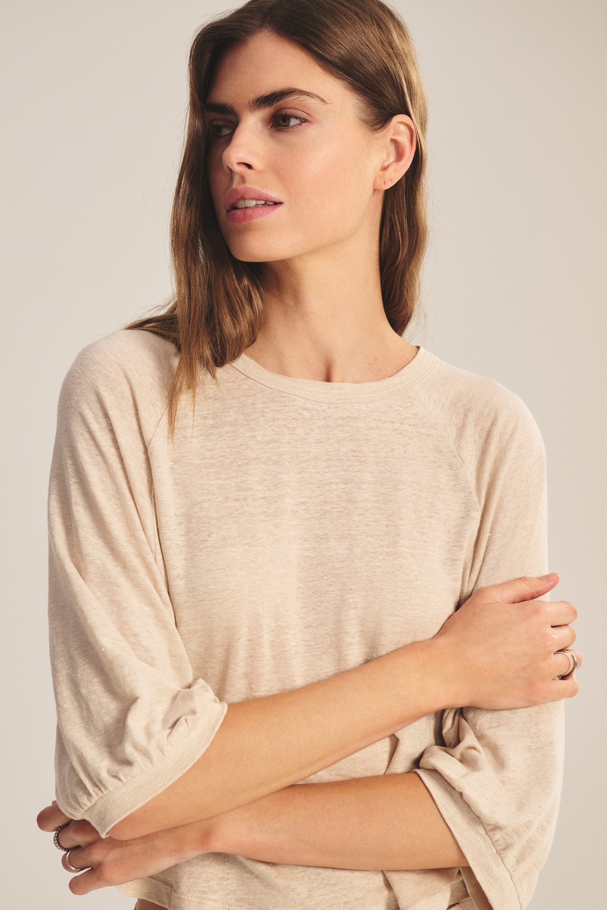 A woman with long brown hair wears the SHELLY TEE by Velvet by Graham & Spencer— a breathable beige linen knit long-sleeve shirt—standing with arms crossed and looking left against a neutral background.-40910148239553