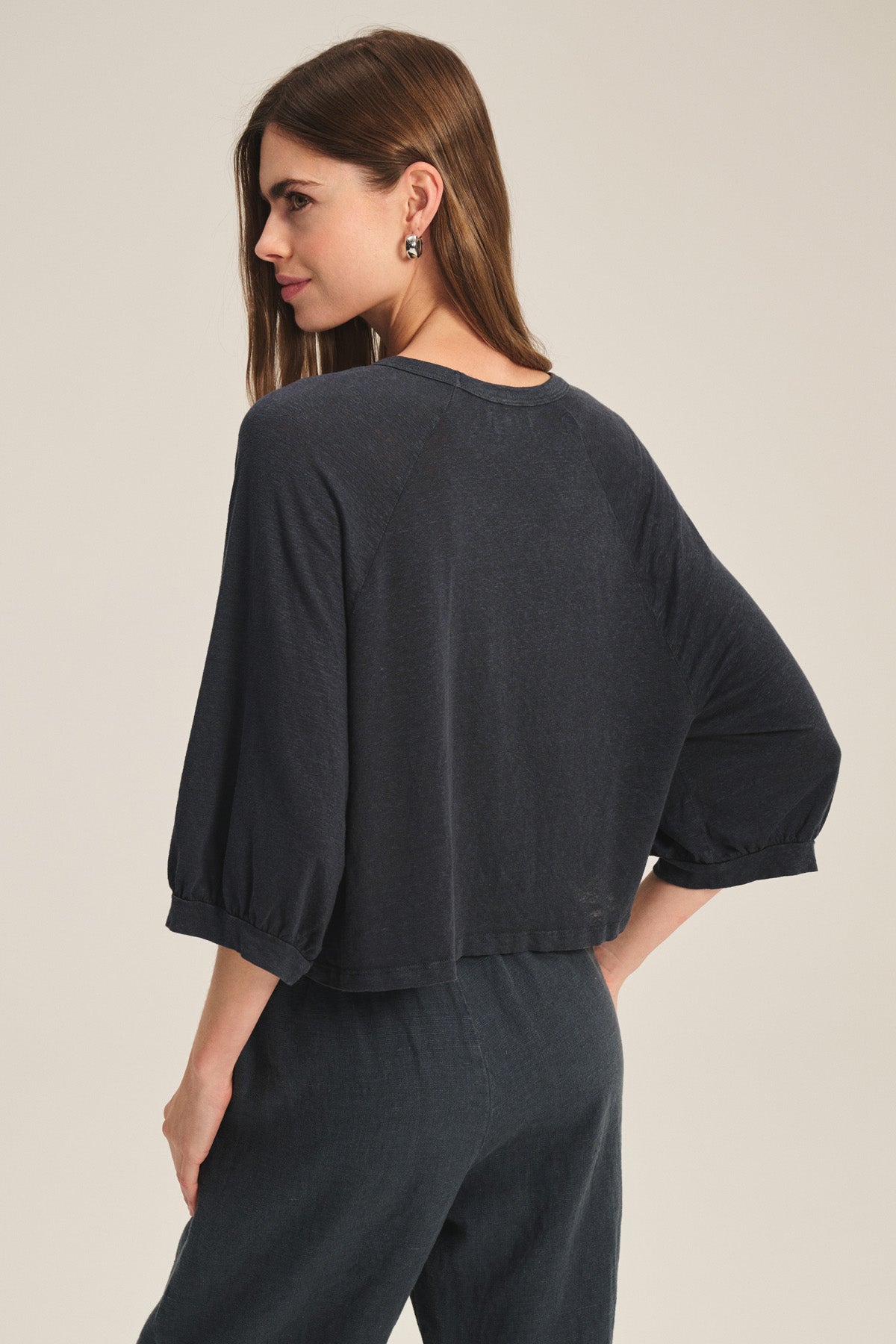 A woman faces away wearing the Velvet by Graham & Spencer SHELLY TEE, a breathable linen knit top with three-quarter sleeves, paired with cropped dark pants against a plain light background.