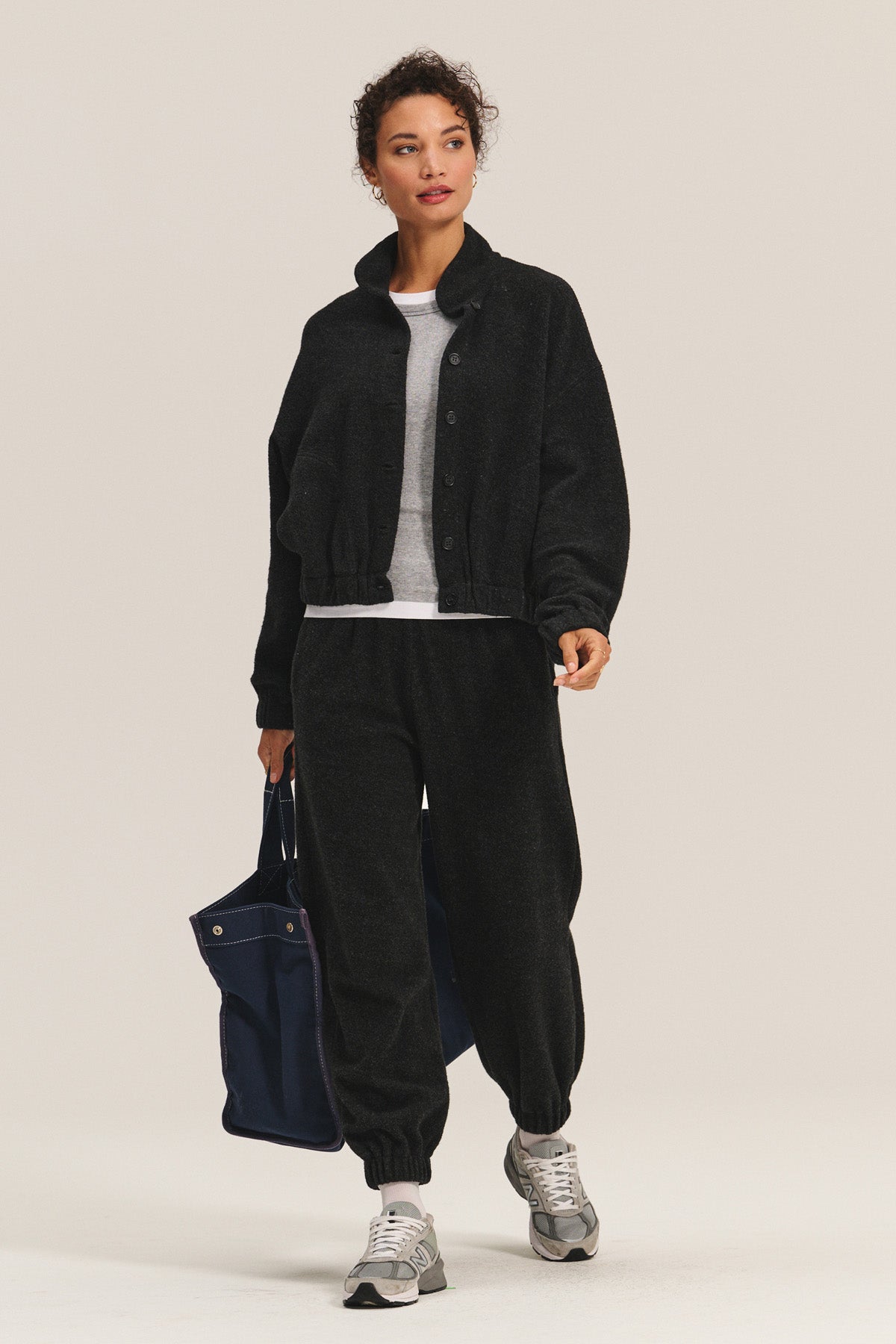 A person wears the Velvet by Graham & Spencer BRIAR JACKET in black sherpa fleece, paired with matching pants, a white shirt, gray sneakers, and a navy tote bag, standing against a plain background.