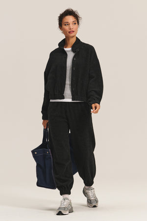 A person wears the Velvet by Graham & Spencer BRIAR JACKET in black sherpa fleece, paired with matching pants, a white shirt, gray sneakers, and a navy tote bag, standing against a plain background.