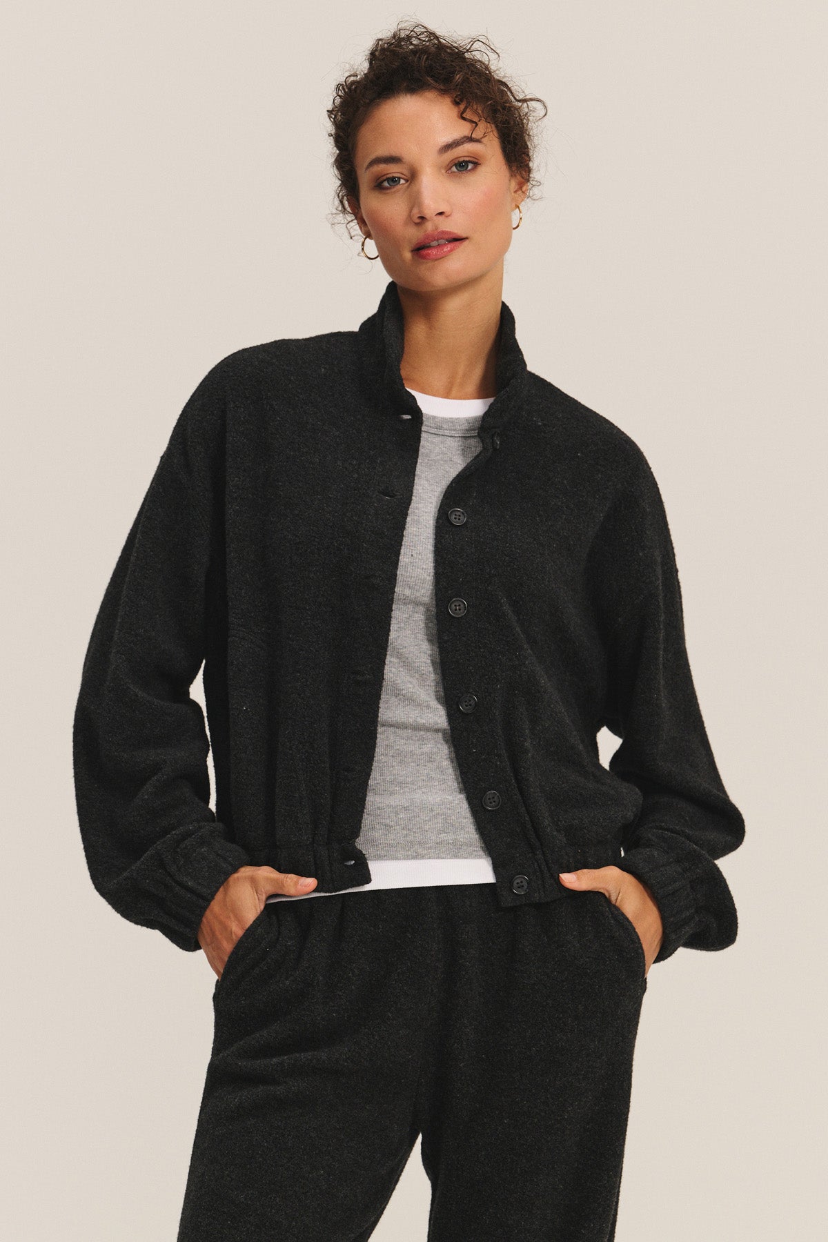 A woman wears the BRIAR JACKET by Velvet by Graham & Spencer—a black, structured cardigan—over a gray T-shirt and matching black pants, standing with hands in pockets against a plain background.-40334112882881