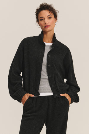 A woman wears the BRIAR JACKET by Velvet by Graham & Spencer—a black, structured cardigan—over a gray T-shirt and matching black pants, standing with hands in pockets against a plain background.