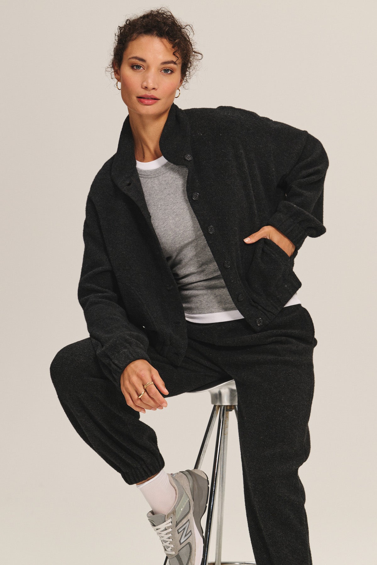 A woman wears the Velvet by Graham & Spencer BRIAR JACKET—a black, flattering button-up—paired with a gray sweatshirt, black pants, and gray sneakers, seated on a stool against a plain light background.-40334112915649