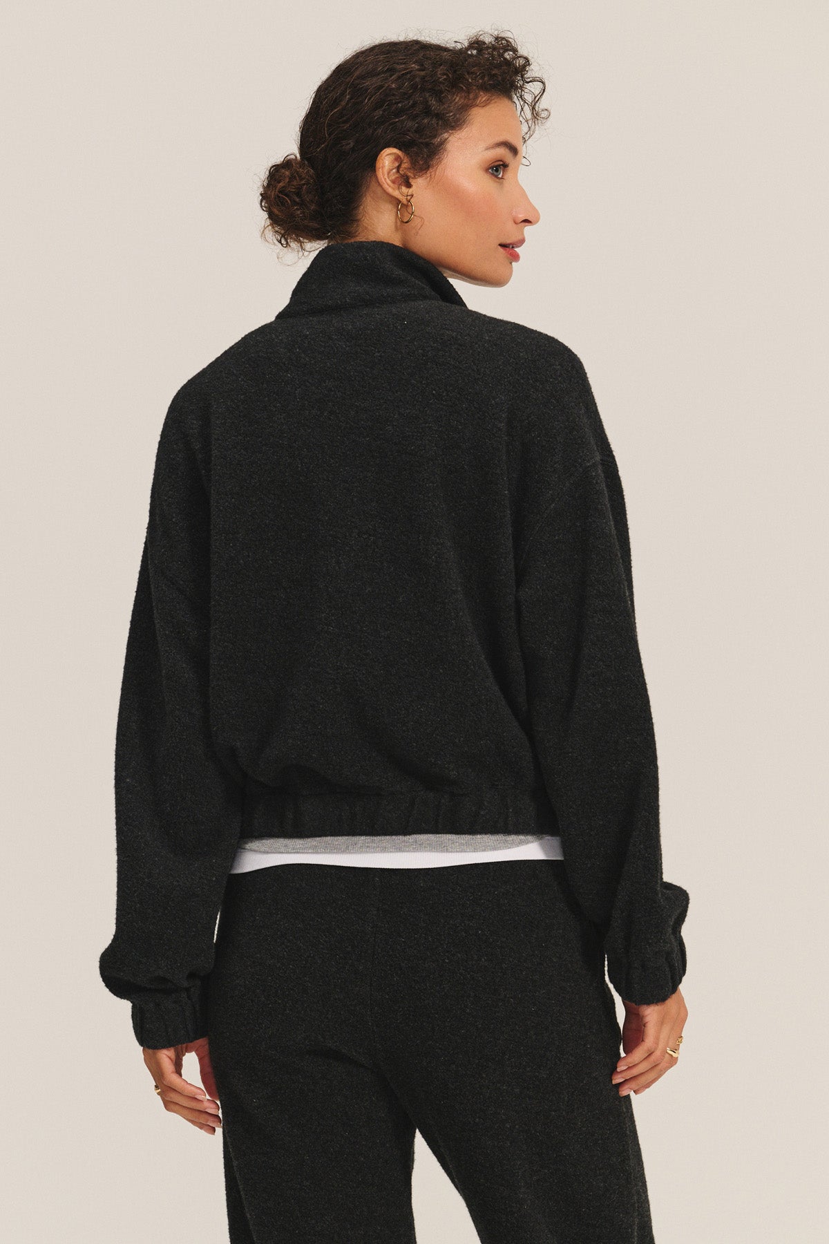 A woman stands with her back to the camera, wearing the Velvet by Graham & Spencer BRIAR JACKET in sherpa fleece, paired with matching black pants and a white shirt visible underneath.