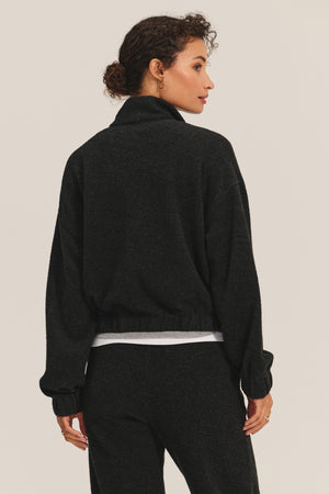 A woman stands with her back to the camera, wearing the Velvet by Graham & Spencer BRIAR JACKET in sherpa fleece, paired with matching black pants and a white shirt visible underneath.