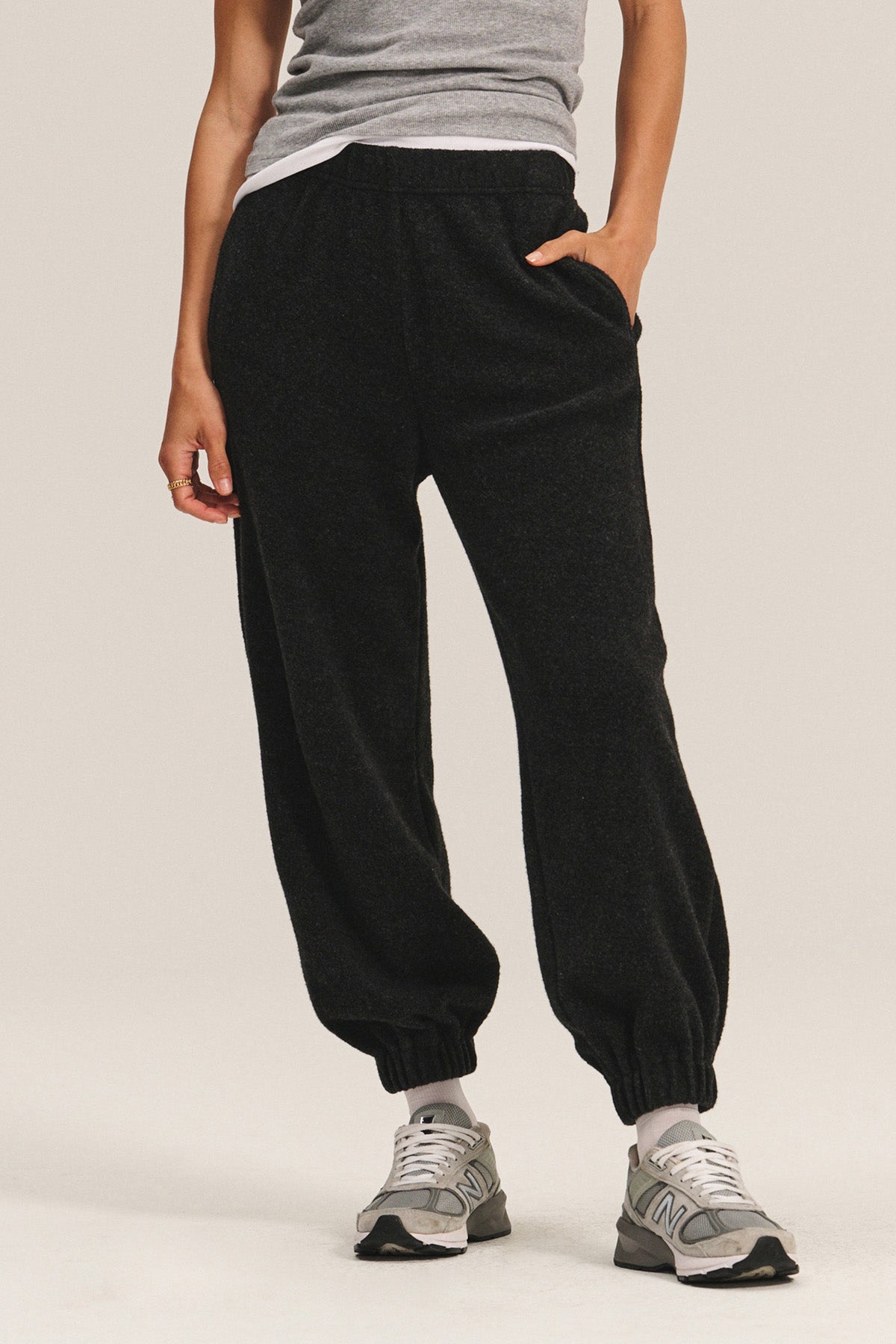 A person wears the DAWN JOGGER sweatpants by Velvet by Graham & Spencer in black with a relaxed silhouette, paired with a grey tank top and grey sneakers, standing against a plain background with one hand in their pocket.-40334159413441