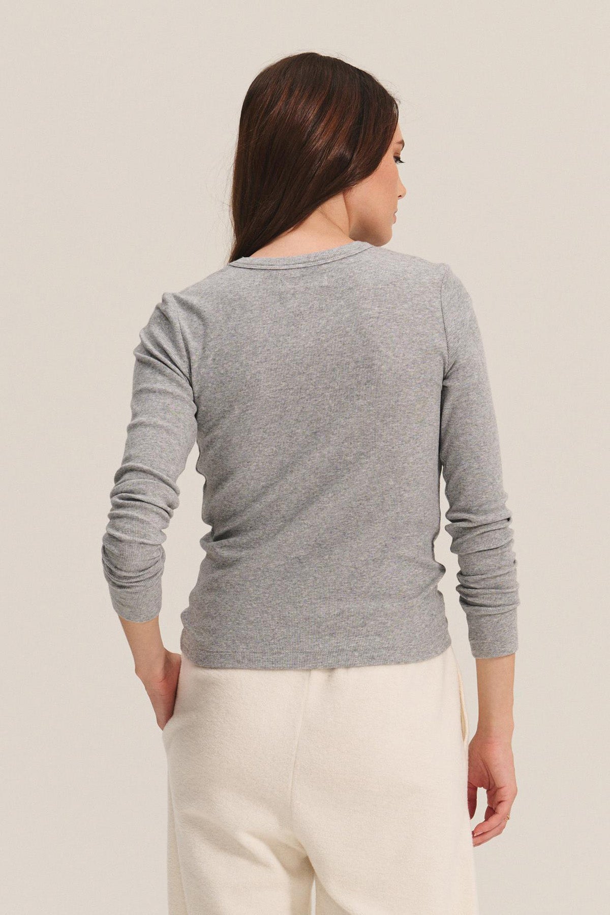 A woman with long brown hair stands facing away, wearing the Velvet by Jenny Graham CAMINO TEE in gray modal rib and off-white pants against a light background—perfect for versatile layering.