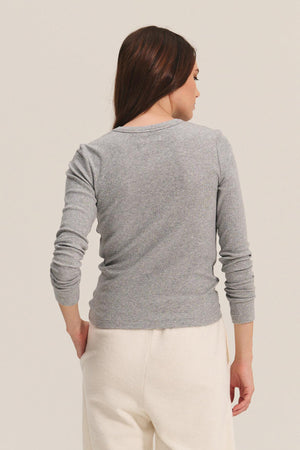 A woman with long brown hair stands facing away, wearing the Velvet by Jenny Graham CAMINO TEE in gray modal rib and off-white pants against a light background—perfect for versatile layering.