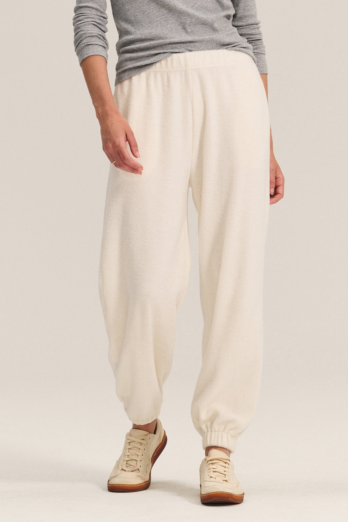 A person stands against a plain background wearing Velvet by Graham & Spencer’s DAWN JOGGER in off-white sherpa fleece with elastic cuffs, paired with a gray long-sleeve top and beige sneakers for a comfortable yet stylish look.