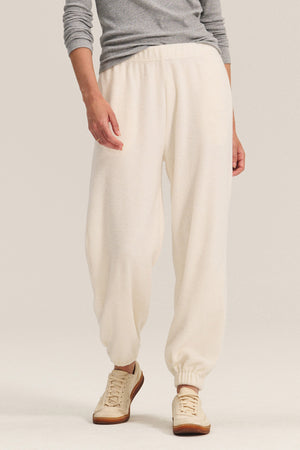 A person stands against a plain background wearing Velvet by Graham & Spencer’s DAWN JOGGER in off-white sherpa fleece with elastic cuffs, paired with a gray long-sleeve top and beige sneakers for a comfortable yet stylish look.