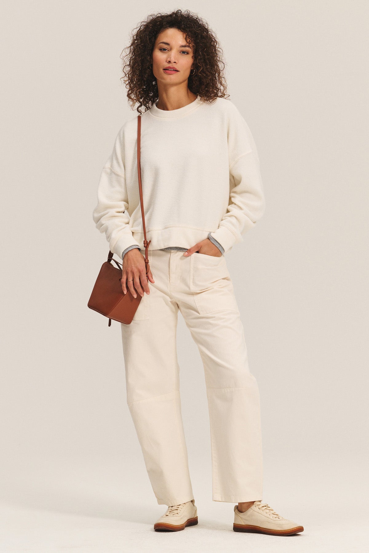 A person with curly hair wears the KIMMY SWEATSHIRT by Velvet by Graham & Spencer, paired with matching pants, beige shoes, and a brown crossbody bag, standing against a plain background.