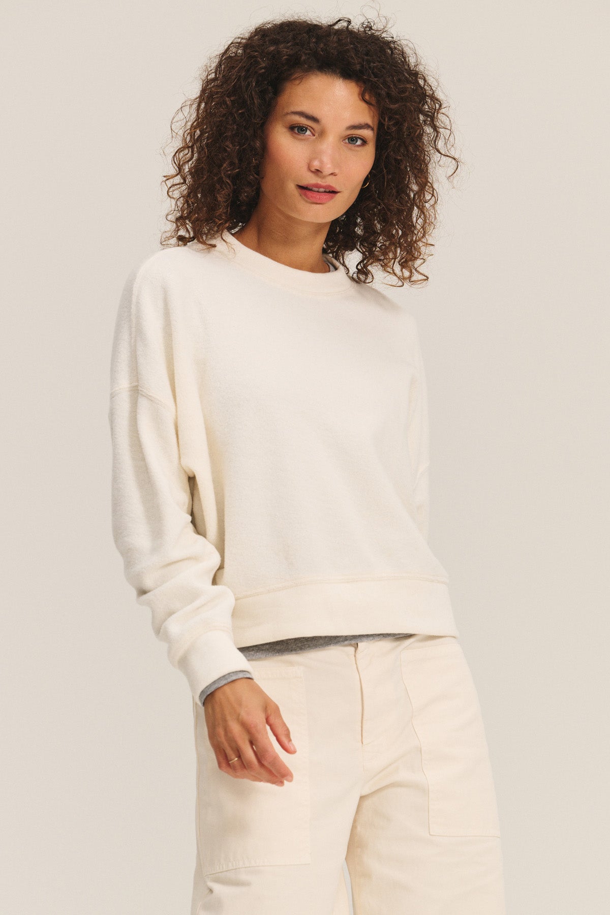 A woman with curly hair wears the KIMMY SWEATSHIRT by Velvet by Graham & Spencer, a cozy sherpa fleece top, paired with light-colored pants against a plain background.
