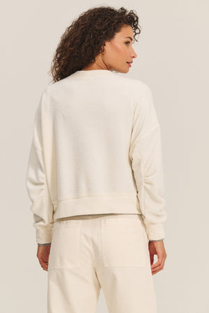 A woman with curly hair wears the Velvet by Graham & Spencer KIMMY SWEATSHIRT and matching pants, facing away from the camera against a neutral background.