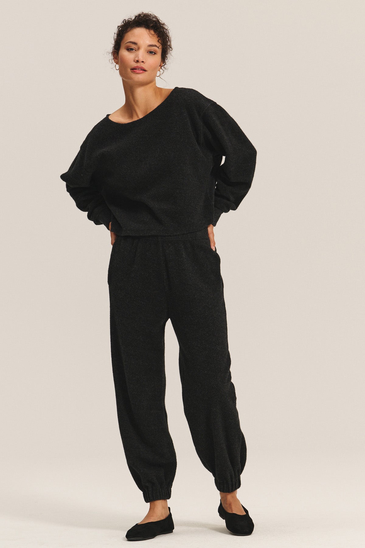 A person stands against a plain background wearing Velvet by Graham & Spencer’s DAWN JOGGER set—loose-fitting black long-sleeve top and matching relaxed jogger pants—styled with black flats.-40334159675585